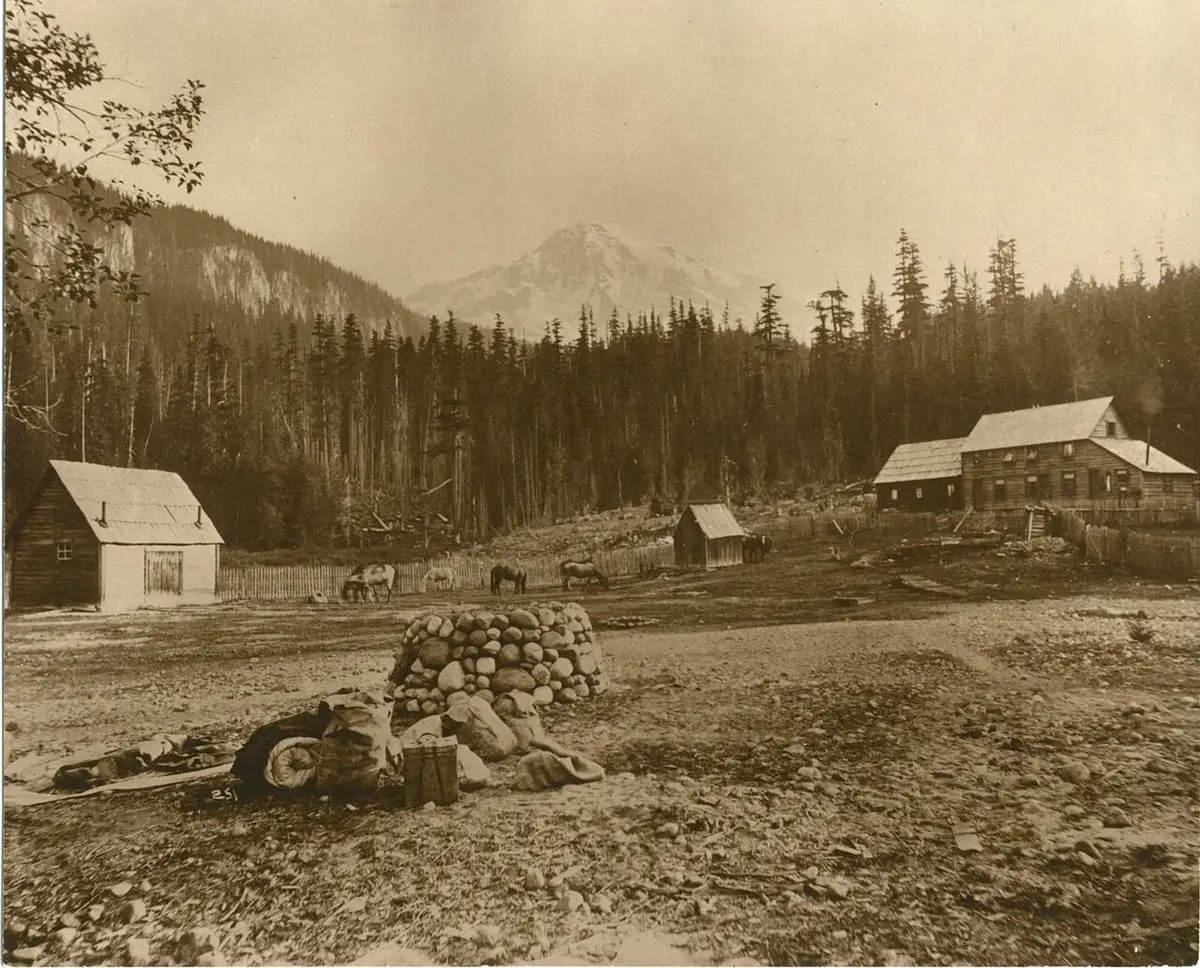 Early sepia view of the Longmire Mineral Springs homestead, with log buildings, grazing horses and Mount Rainier beyond