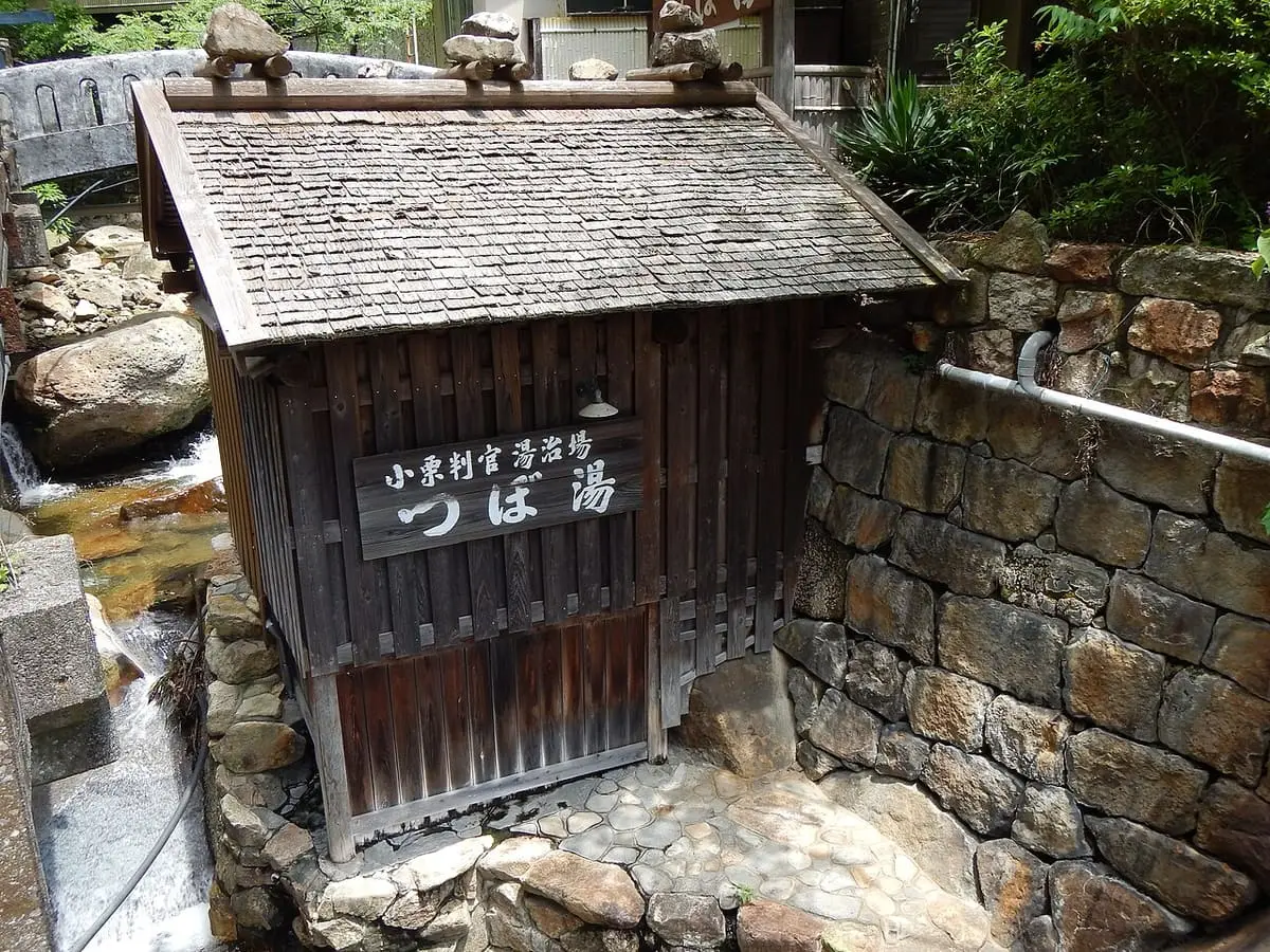 Small wooden Tsuboyu bath hut with shingle roof beside a stone wall in Yunomine