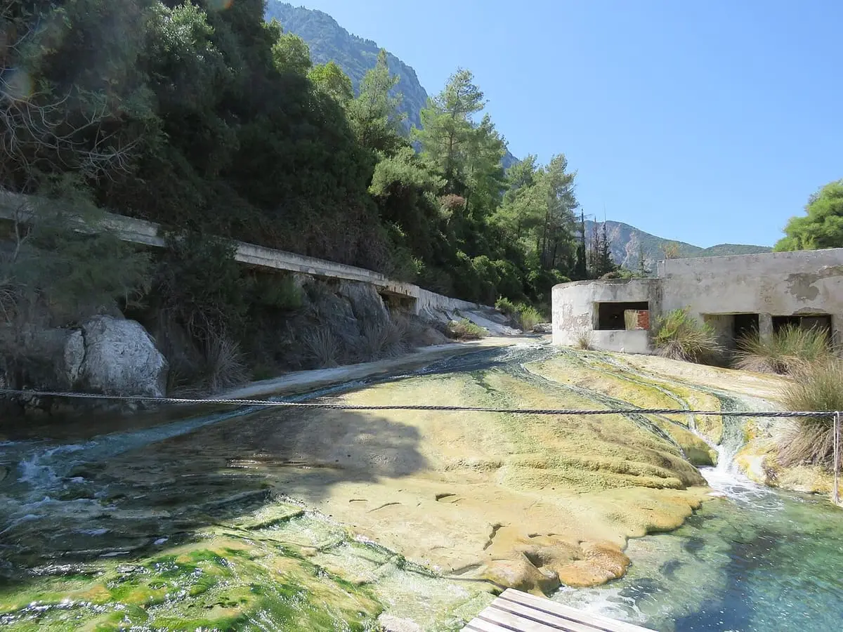 Mineral stained travertine terraces with shallow hot water below wooded hills at Thermopylae