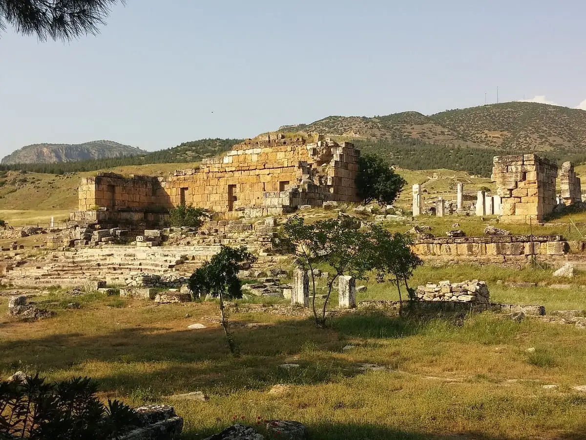 Ruined stone buildings of ancient Hierapolis in a grassy field below green hills