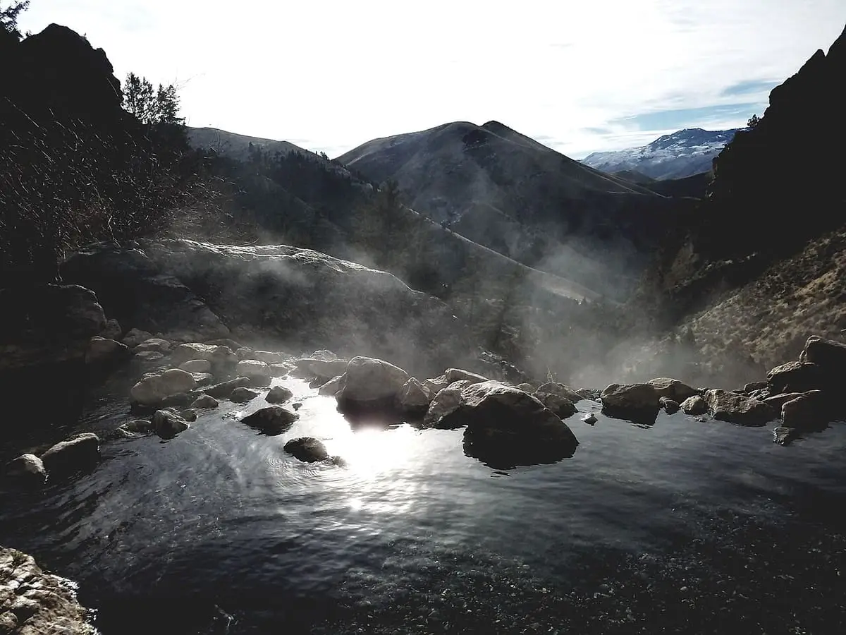 Steam rising off a boulder-dammed hot pool at Goldbug Hot Springs with hills and snowy peaks beyond
