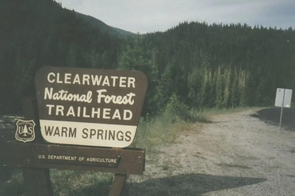 Wooden Clearwater National Forest sign for the Warm Springs Trailhead beside a gravel road and forested hills