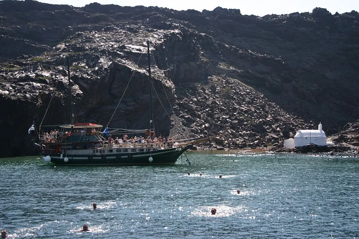 Tour boat anchored in the Palea Kameni hot springs bay below dark volcanic cliffs