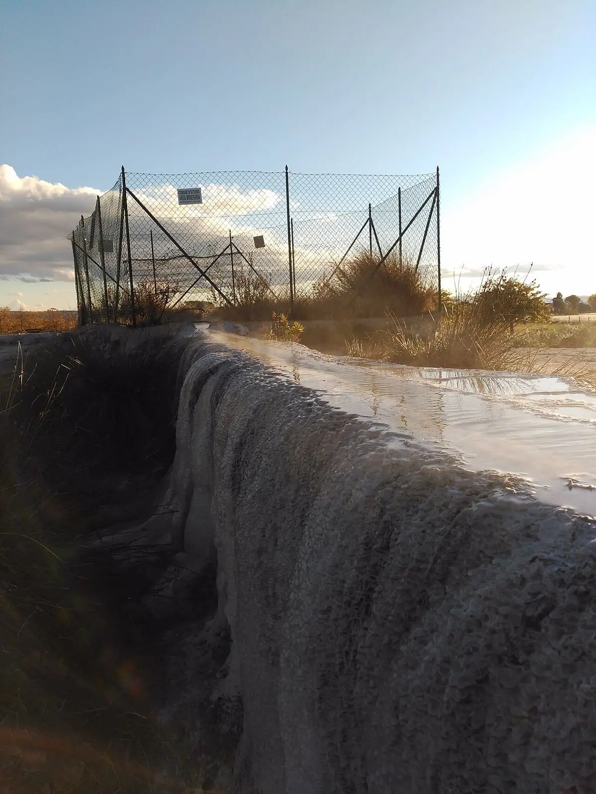 Hot sulphur water spilling down a travertine bank beside a fenced channel at Piscine Carletti