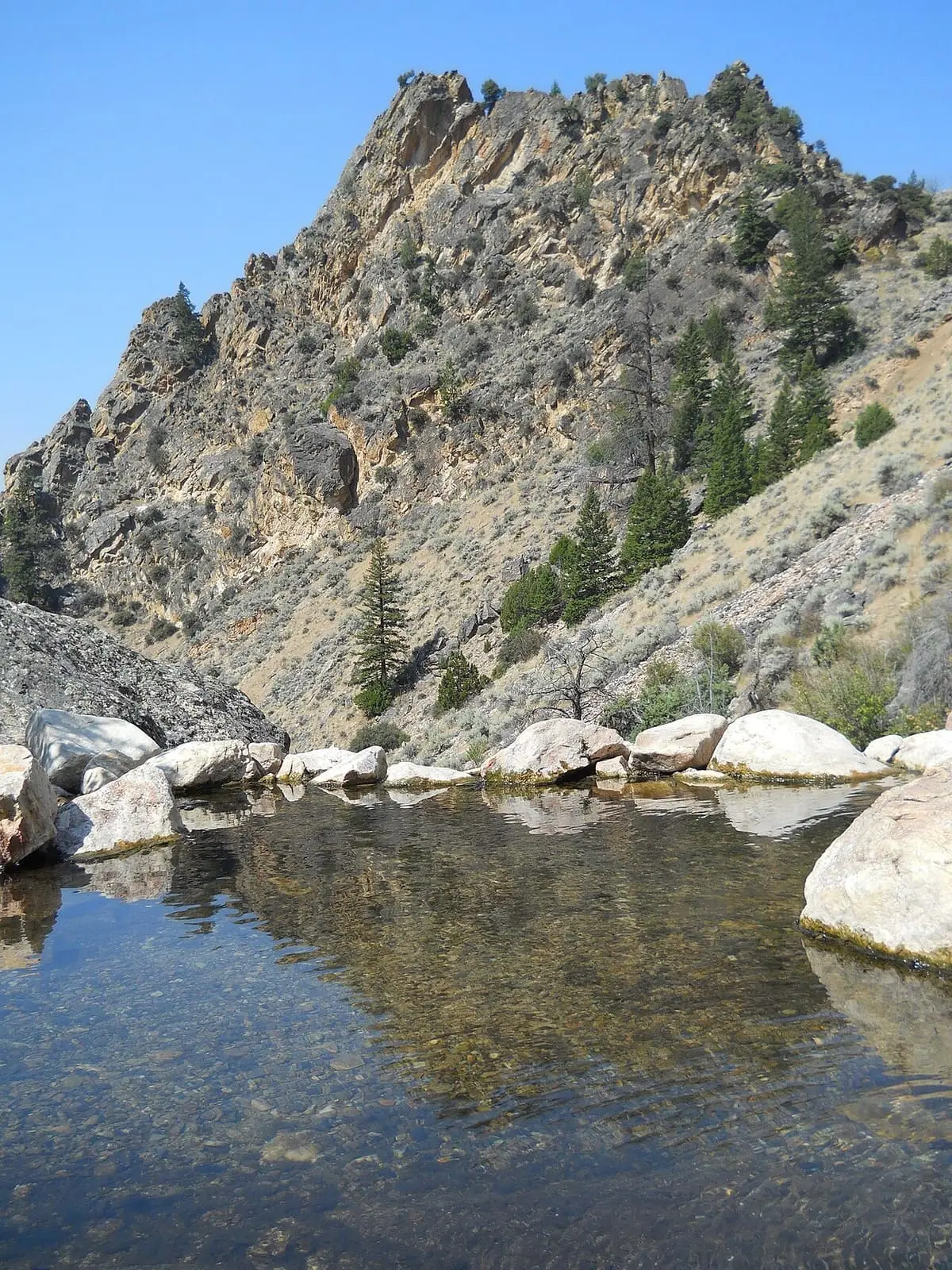 Clear soaking pool ringed by pale boulders below a craggy canyon wall at Goldbug Hot Springs