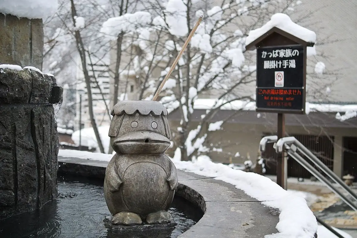 Snow covered kappa statue beside a hot spring hand bath and wooden sign in Jozankei