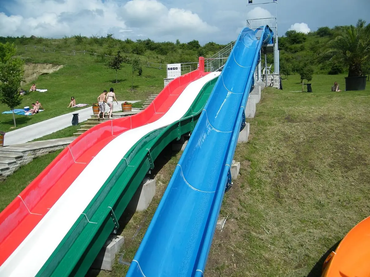 Red, white and green outdoor slide beside a blue slide on a grassy hillside at Demjén