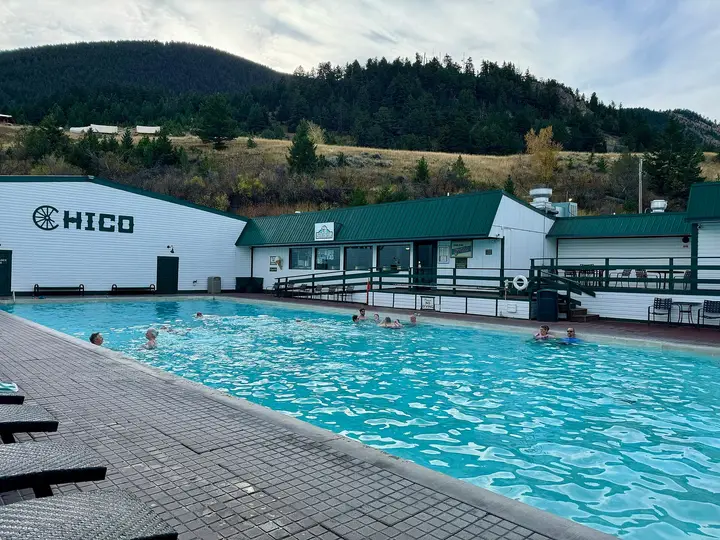 Large outdoor hot pool at Chico Hot Springs below white buildings with the Chico name and forested hills