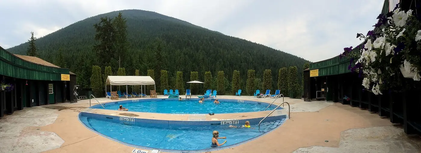 Outdoor hot pool at Nakusp Hot Springs surrounded by green buildings and a forested hill