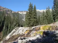Mineral deposit mound streaked with yellow and green at Dewar Creek Hot Springs below forested peaks
