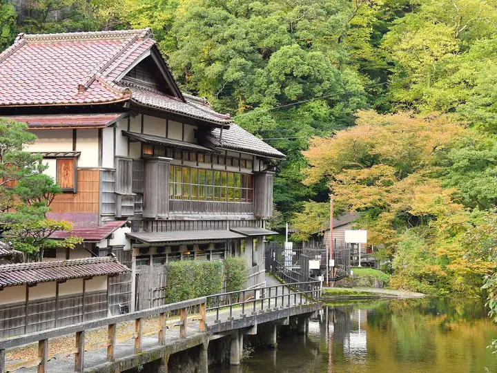 Traditional wooden inn beside a calm river and green hillside at Higashiyama Onsen
