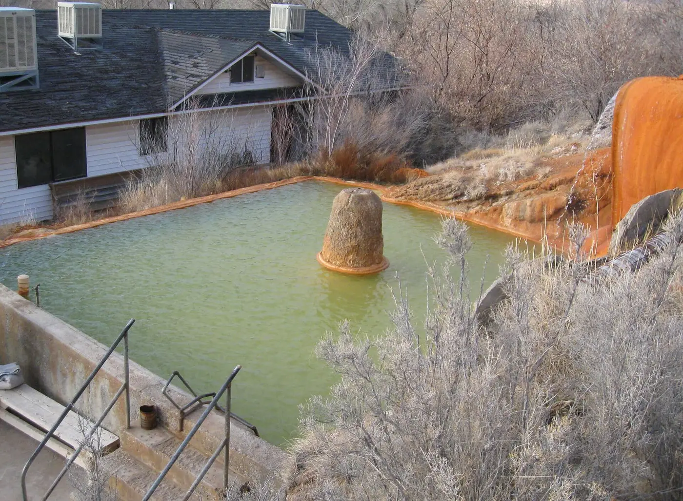 Green mineral pool at Mystic Hot Springs beside orange travertine mounds and frosted brush
