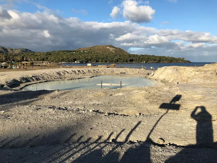 Pale grey mud pool of the Laghetti di Fanghi on Vulcano with wooded hills and the bay behind