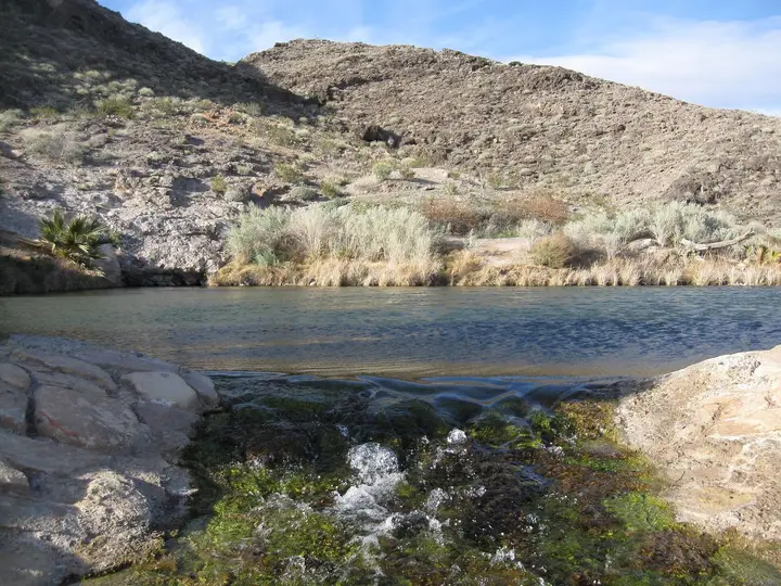 Clear warm spring pool at Rogers Spring edged by rocks, grasses and rocky desert hills