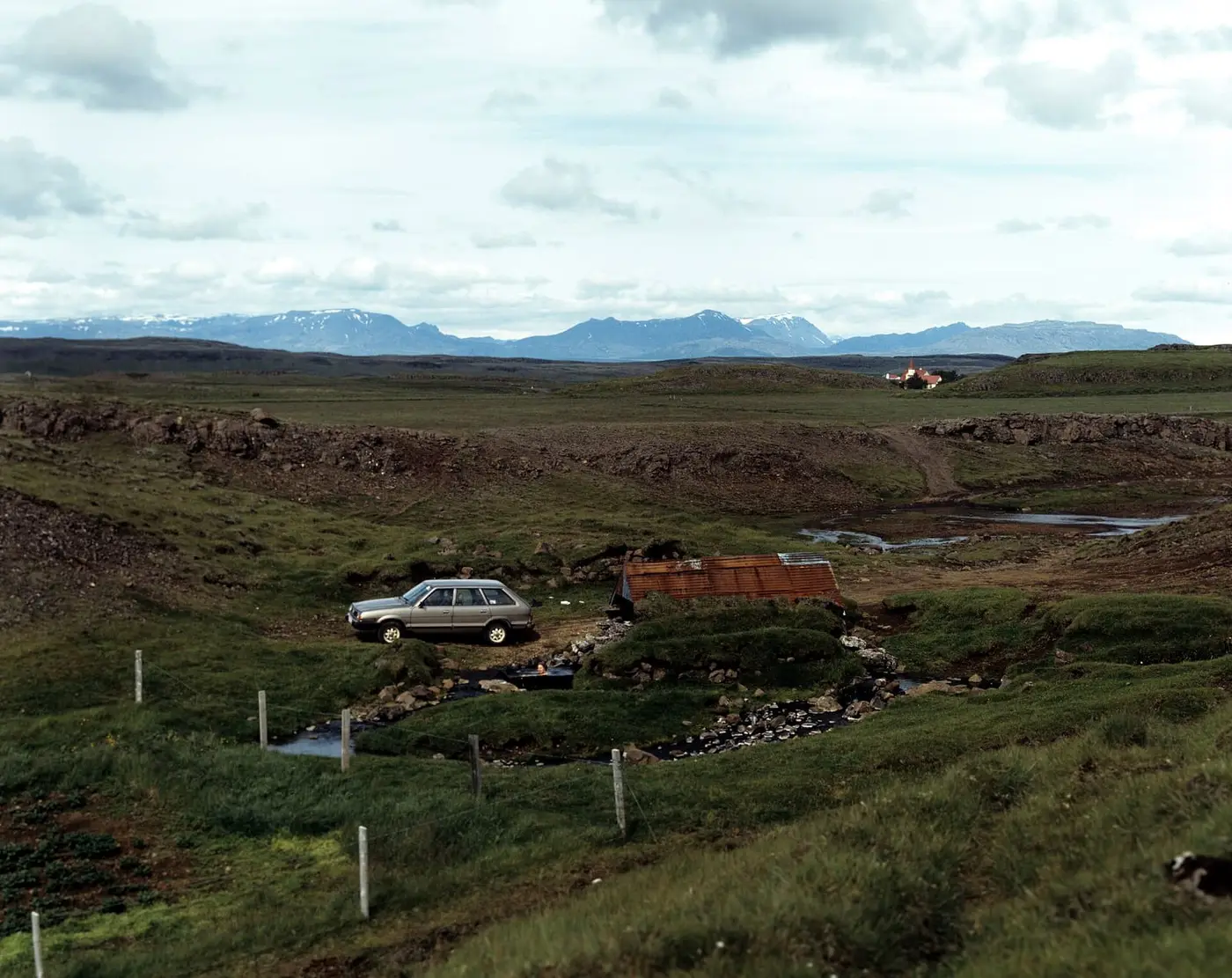 Small rusty roofed hut and warm stream at Hrunalaug with the Hruni farm and mountains beyond