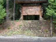 Carved wooden Belknap Hot Springs Resort sign under a small roof in tall forest