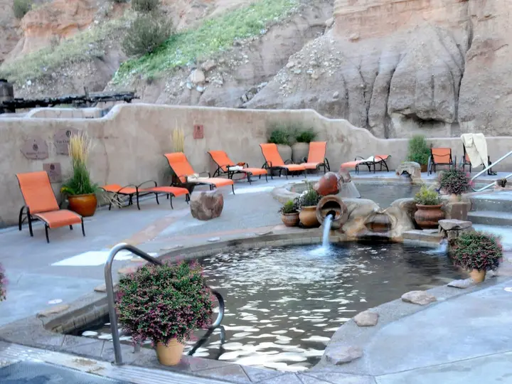 Mineral pool with clay pot fountain and orange lounge chairs below sandstone cliffs at Ojo Caliente