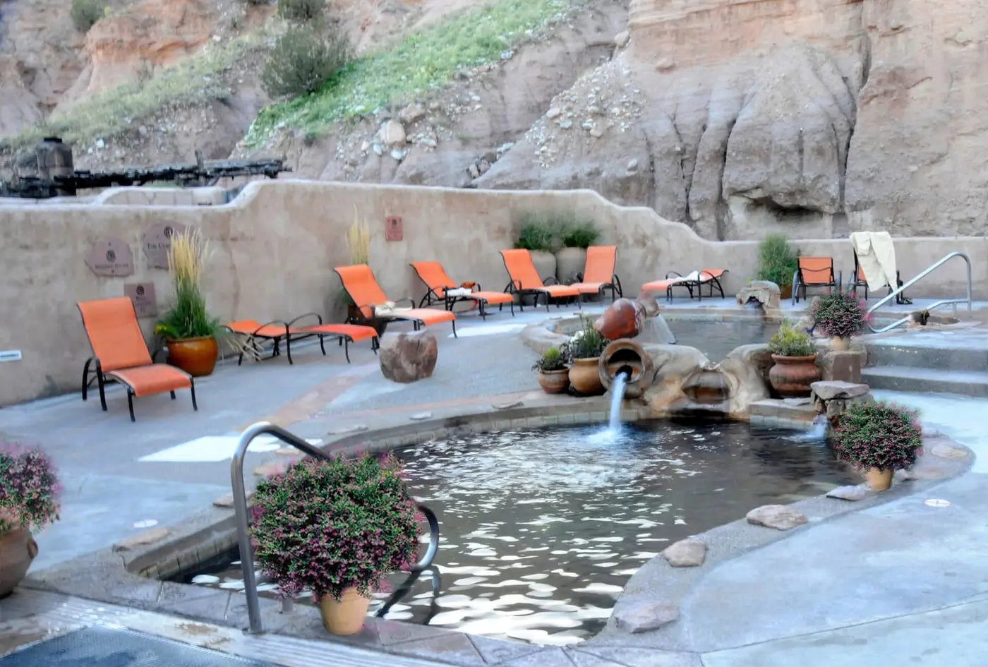 Mineral pool with clay pot fountain and orange lounge chairs below sandstone cliffs at Ojo Caliente