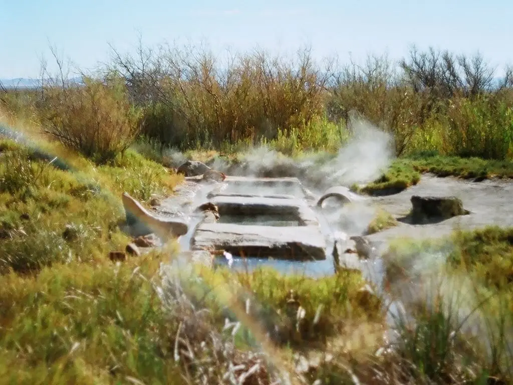 Steaming concrete soaking channels at Baker Hot Springs surrounded by grass and desert brush under blue sky
