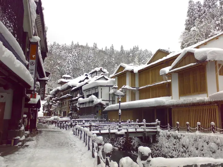 Snow covered wooden inns lining the river in Ginzan Onsen with forested hills behind