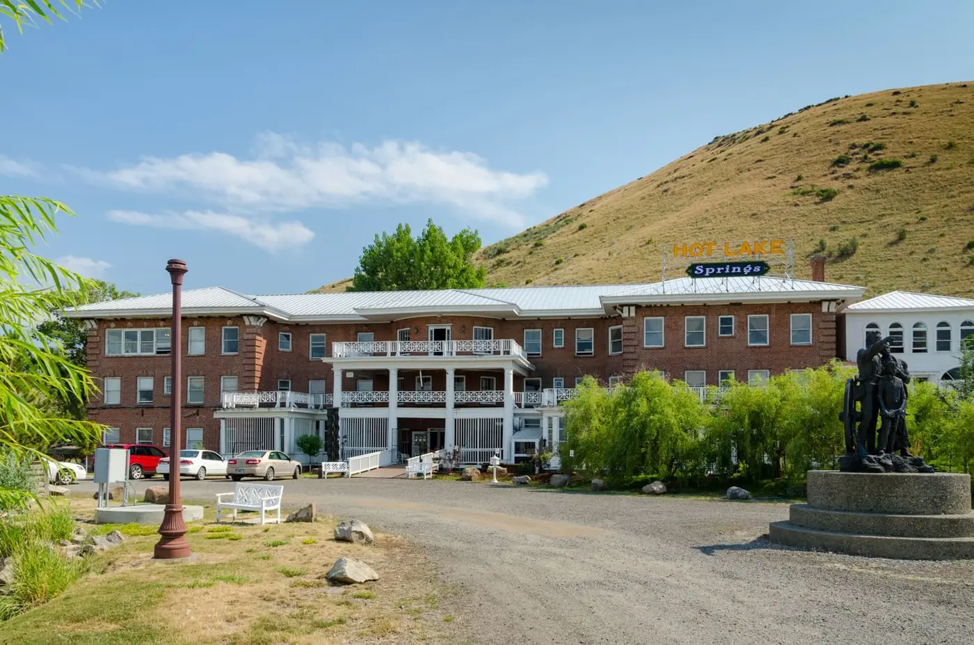 Brick main building of Hot Lake Springs Resort below a dry grassy hillside with a bronze statue out front