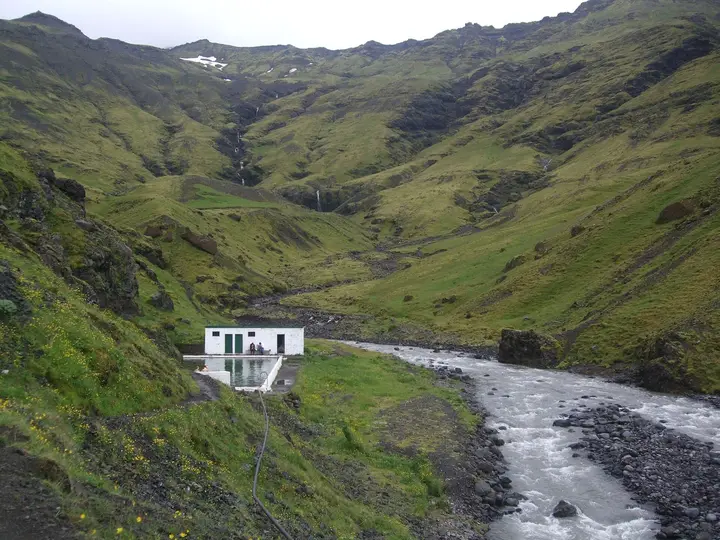 Seljavallalaug pool and white changing house beside a rushing river in a steep green valley