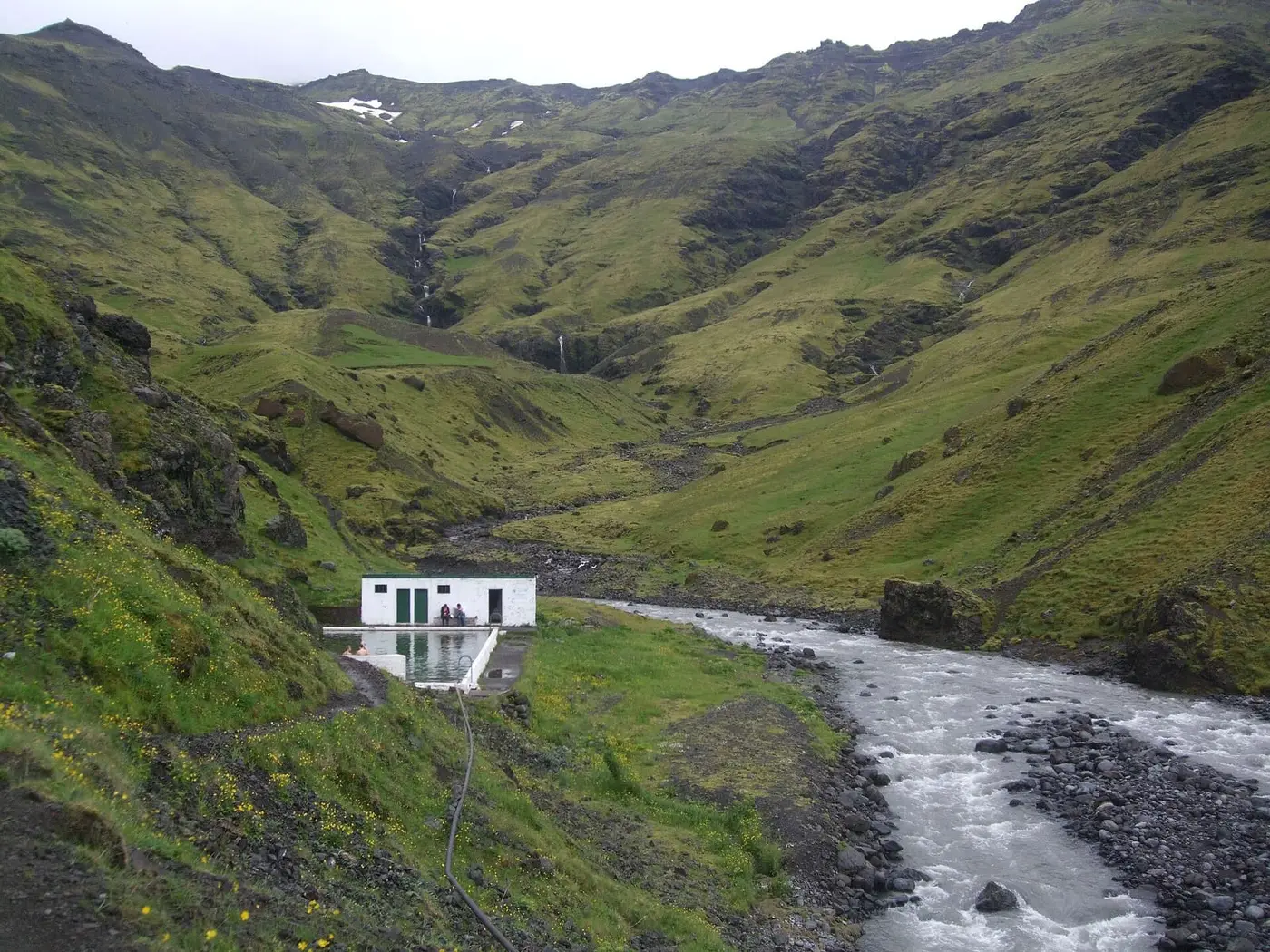 Seljavallalaug pool and white changing house beside a rushing river in a steep green valley