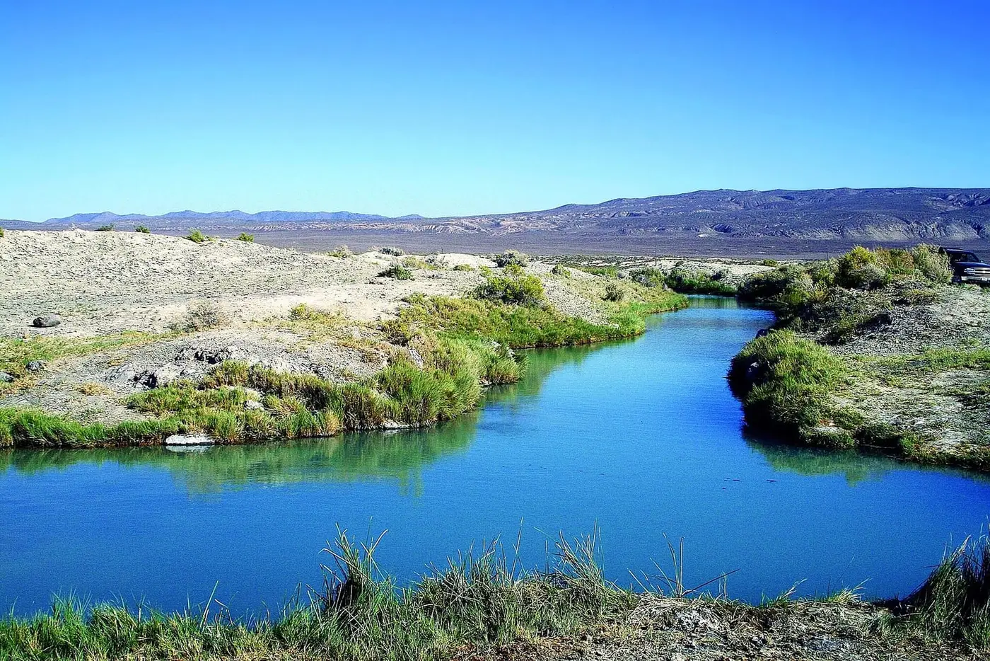 Blue hot water channel at Trego Hot Springs edged with green grass and pale desert ground