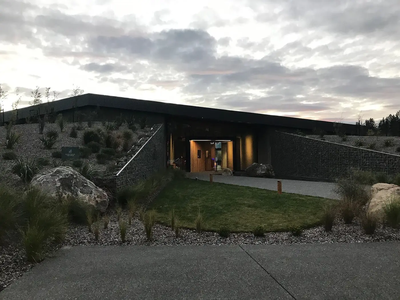 Low modern entrance building of Opuke Thermal Pools with a lit doorway and native plantings at dusk