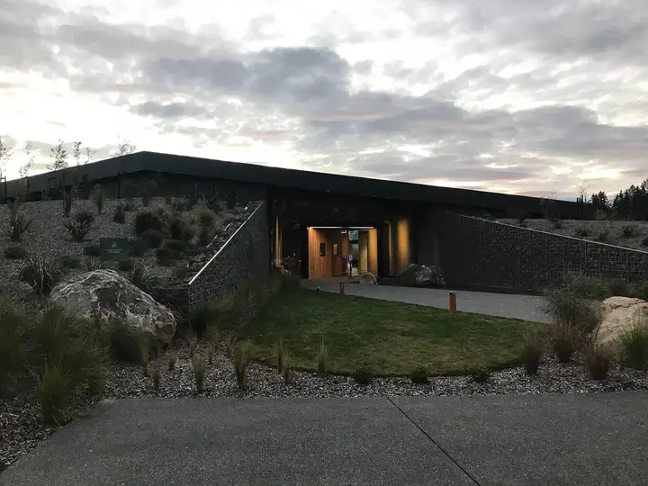 Low modern entrance building of Opuke Thermal Pools with a lit doorway and native plantings at dusk