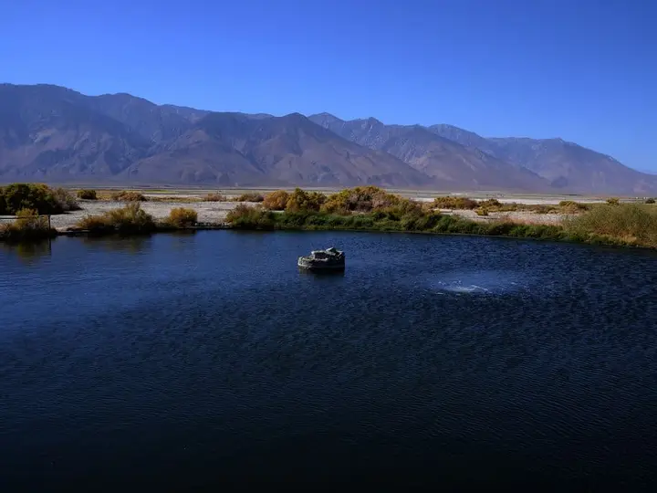 Dark blue pool of Dirty Socks Hot Spring with a concrete wellhead, desert brush and bare mountains beyond