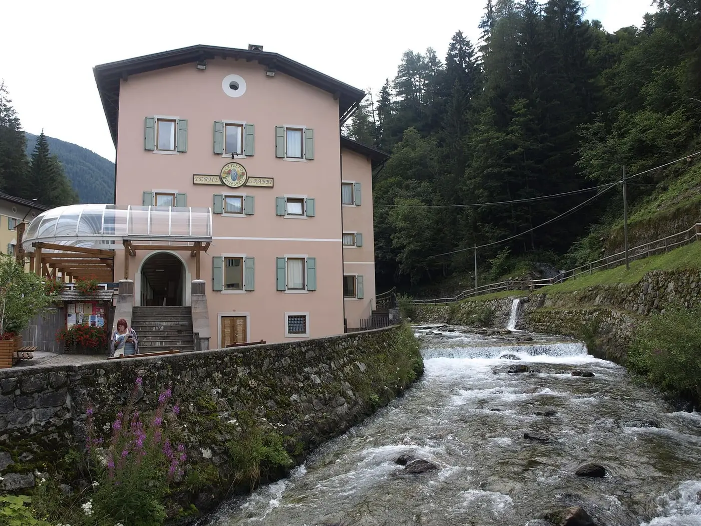 Pink Terme di Rabbi spa building with green shutters beside a rushing mountain stream
