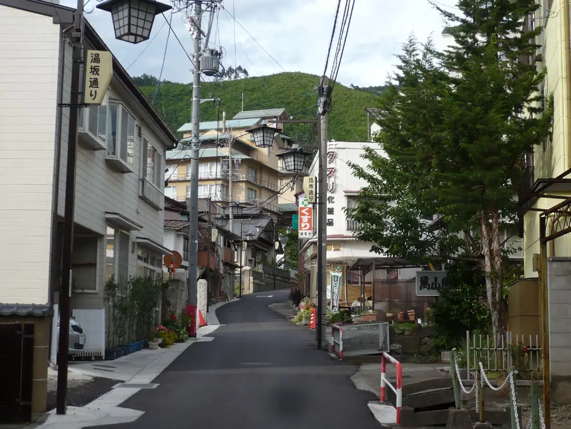 Narrow street climbing through Asama Onsen town past inns toward green hills in Matsumoto