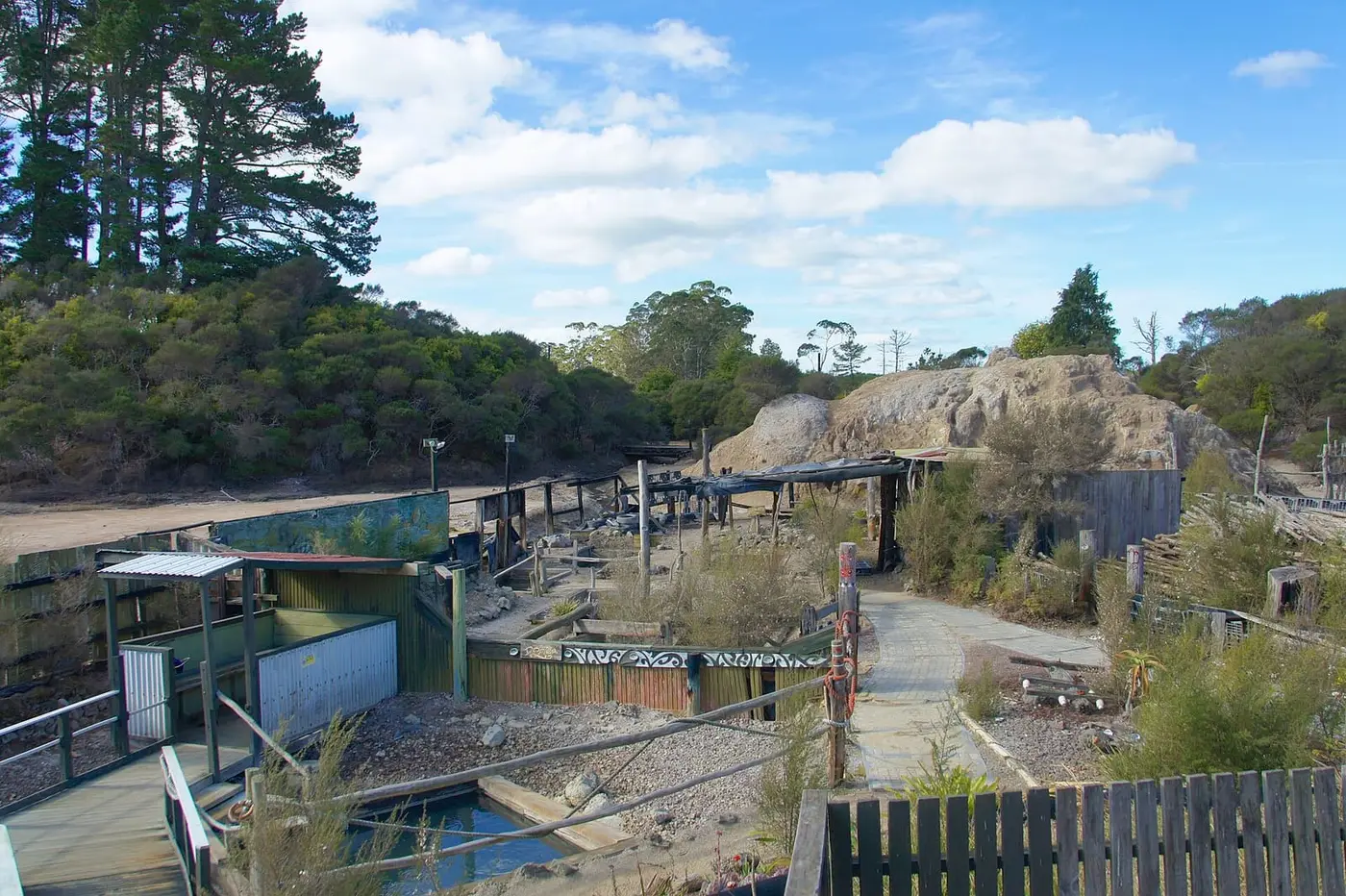 Wooden fenced pool enclosures and walkways of the Ngawha Springs complex under a blue sky