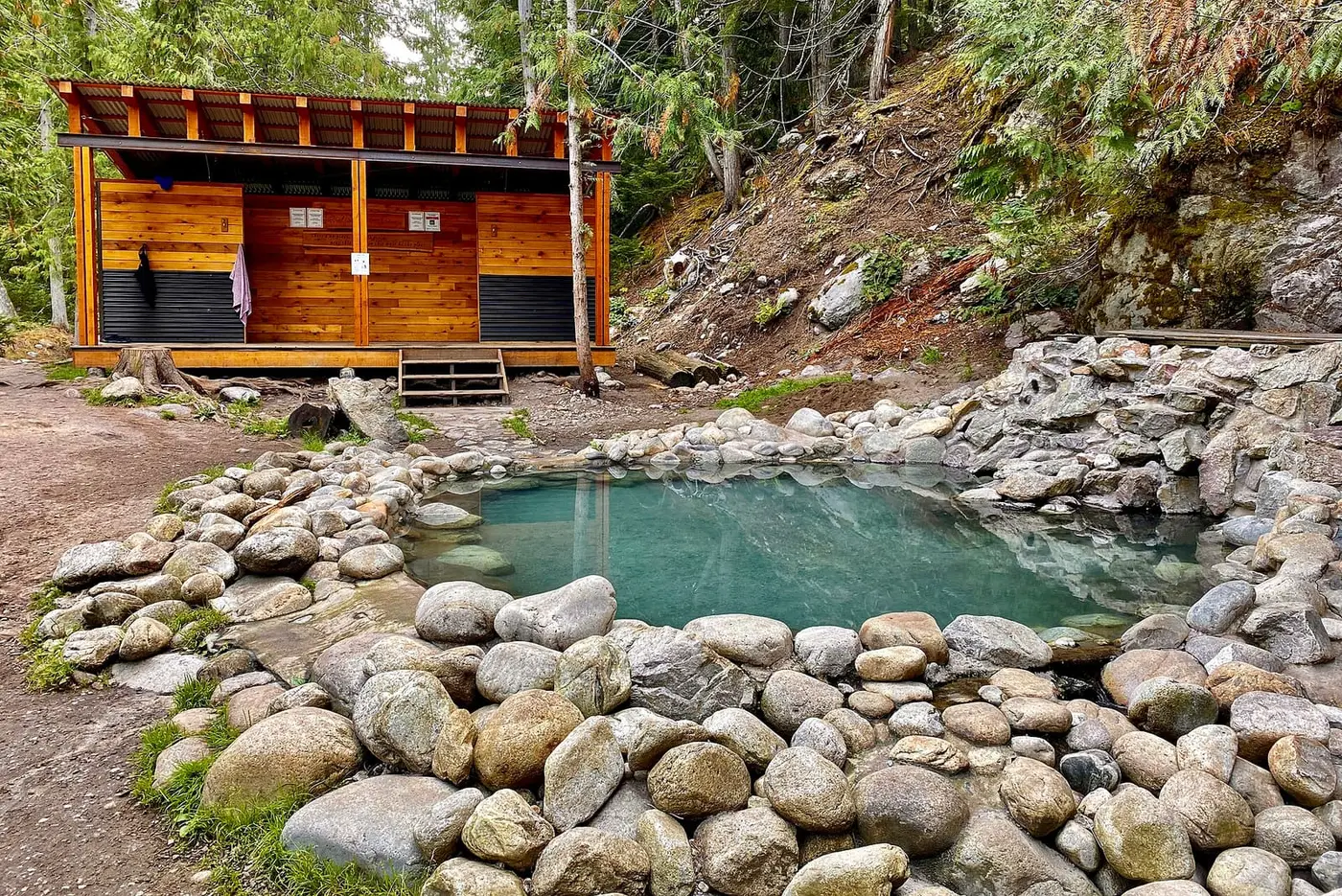 Rock-lined main pool at Halfway River Hot Springs beside a wooden change shelter in the forest