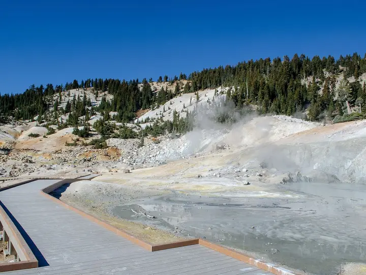 Bumpass Hell boardwalk passing a gray boiling pool with steam rising under a deep blue sky