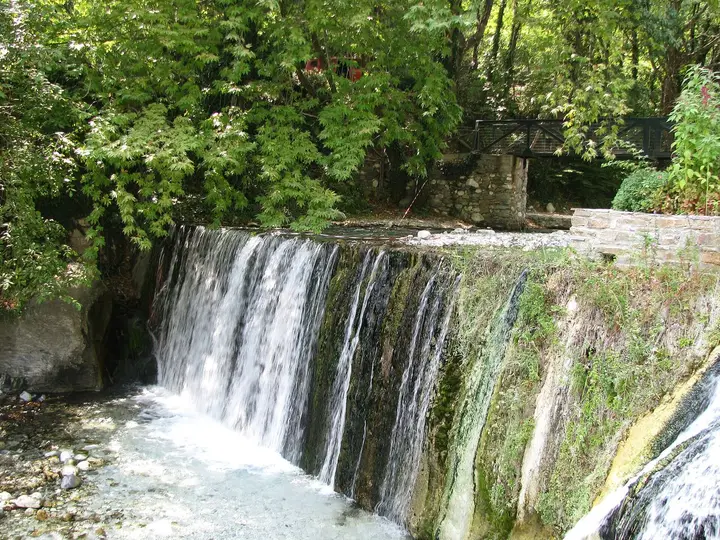 Thermal waterfall spilling over a mossy ledge at Loutra Pozar beneath green trees