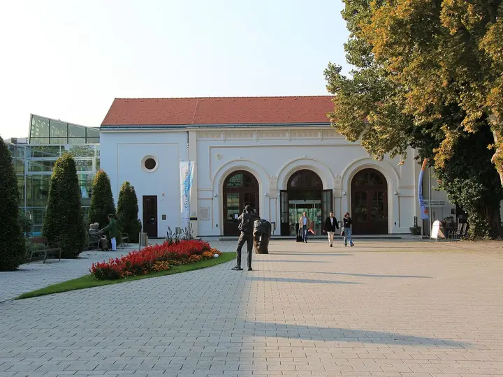Paved forecourt and arched white entrance building of the Römertherme Baden beside its glass hall
