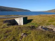 Small stone hot pool and wooden bath hut in the grass beside the road above a fjord