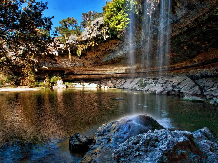 Waterfall pouring from a limestone overhang into the green grotto pool at Hamilton Pool Preserve