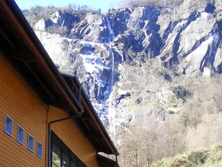 Timber clad Terme di Premia building below a tall waterfall on granite cliffs in Valle Antigorio
