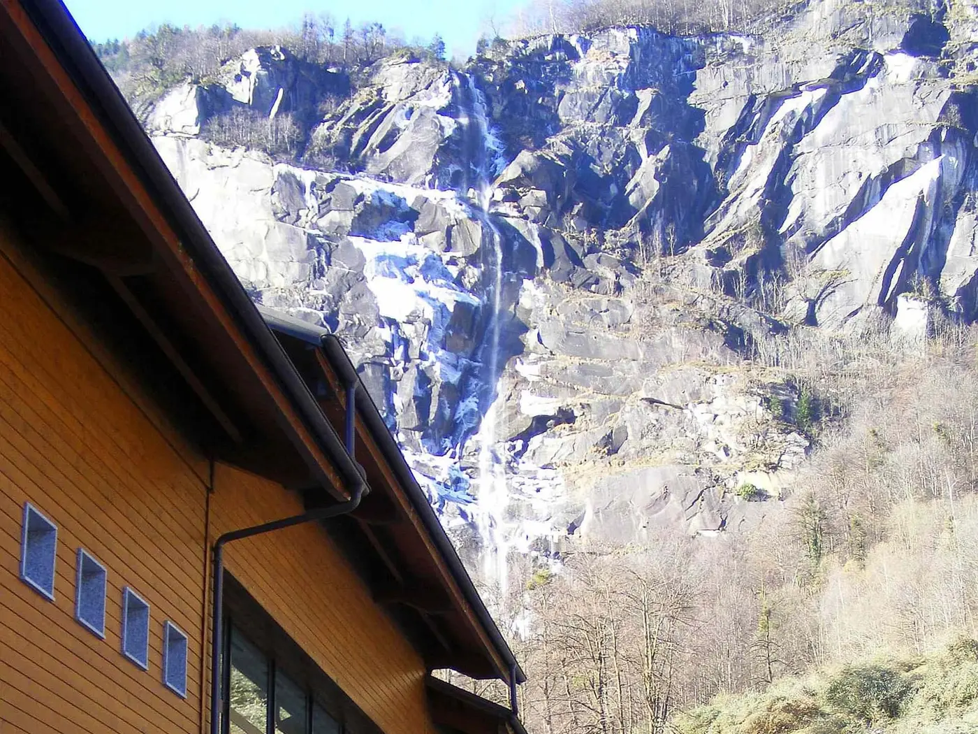Timber clad Terme di Premia building below a tall waterfall on granite cliffs in Valle Antigorio